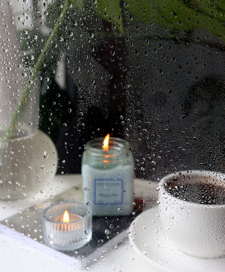Warm candlelight and a cup of coffee by a rain-soaked window create a cozy atmosphere.