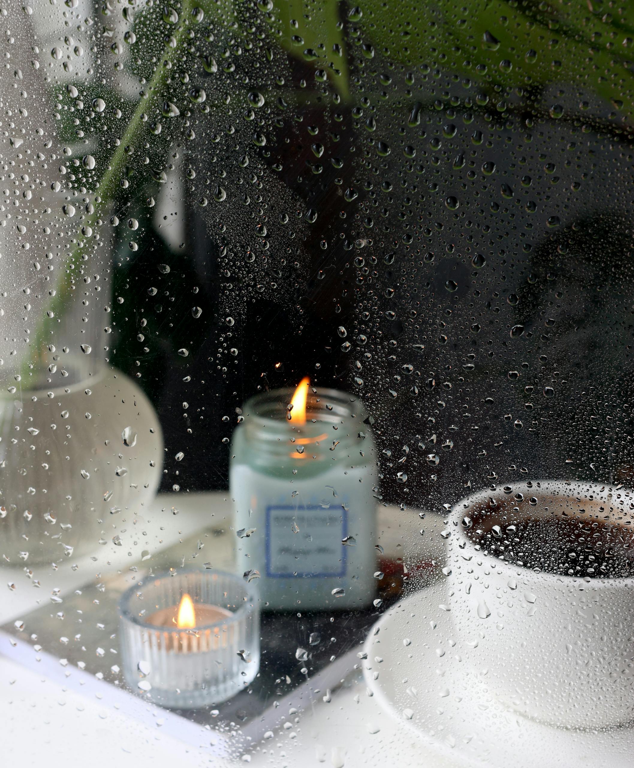 Warm candlelight and a cup of coffee by a rain-soaked window create a cozy atmosphere.